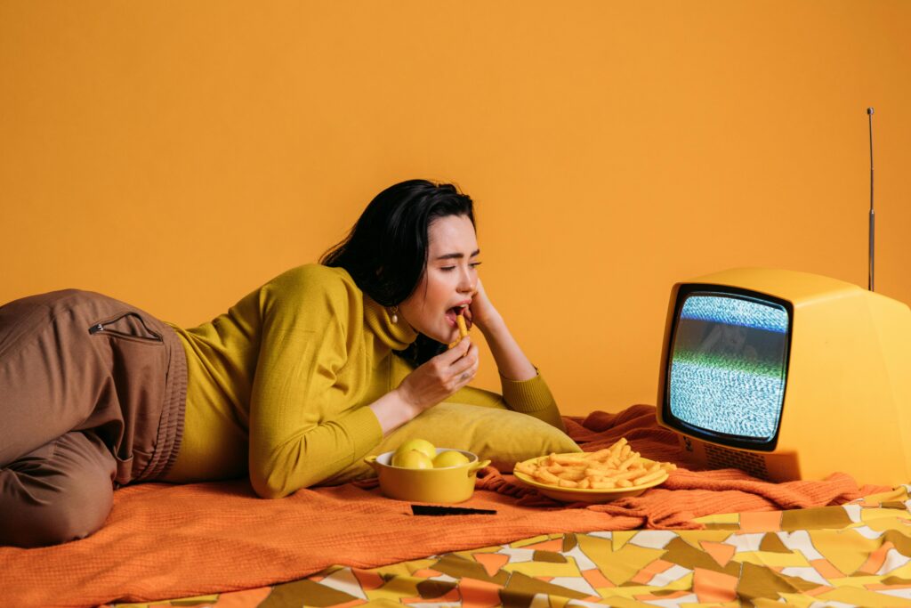 Photo by KoolShooters : https://www.pexels.com/photo/woman-in-yellow-long-sleeve-shirt-watching-tv-and-eatinghi-6977372/