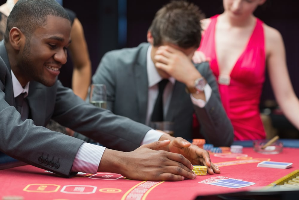 man-collecting-chips-in-casino-with-another-man-holding-his-head-in-hands-in-the-background
