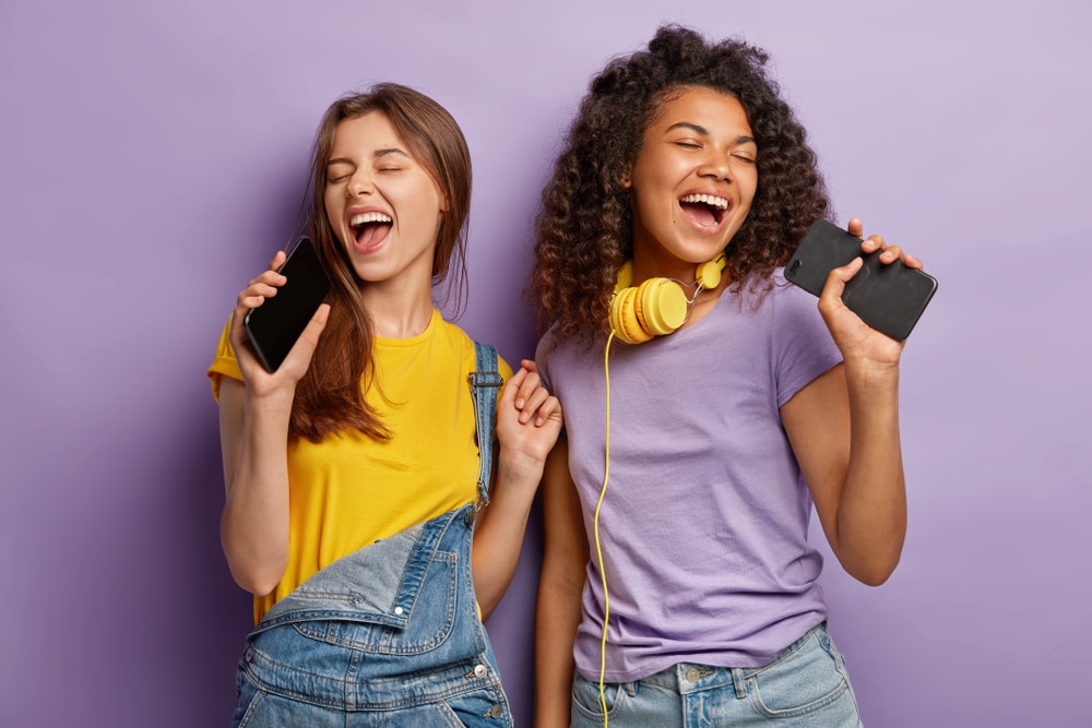 two teenage girls holding their phones and singing against a purple background