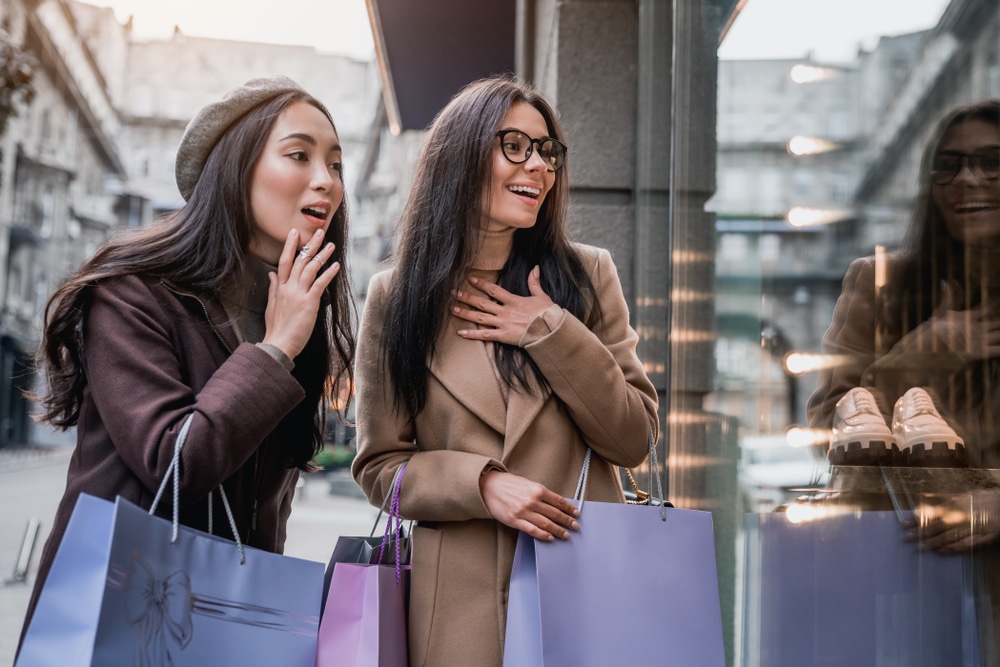 two young brown haired women holding shopping bags looking through a store window