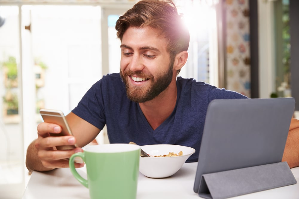 young blonde man eating breakfast while smiling at his phone