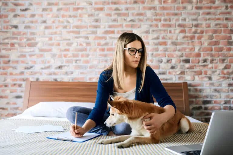 woman and dog on the bed taking notes while looking at her laptop