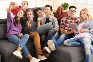 group of teens with modern devices taking a selfie while sitting on a sofa indoors