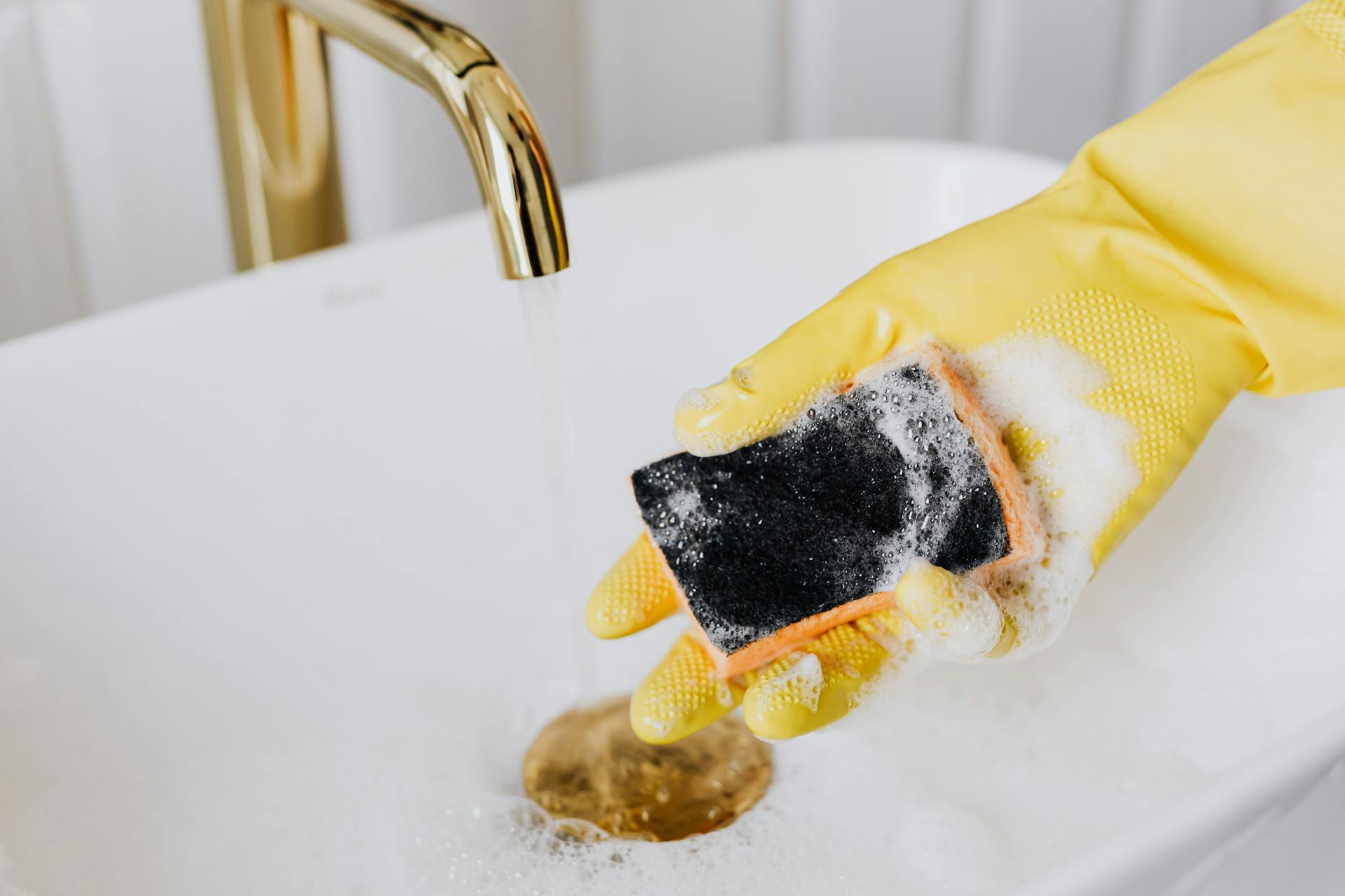 hand-in-yellow-glove-holding-wet-soapy-sponge-over-sink