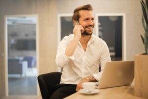 man on the phone smiling with a coffee cup and laptop on the table