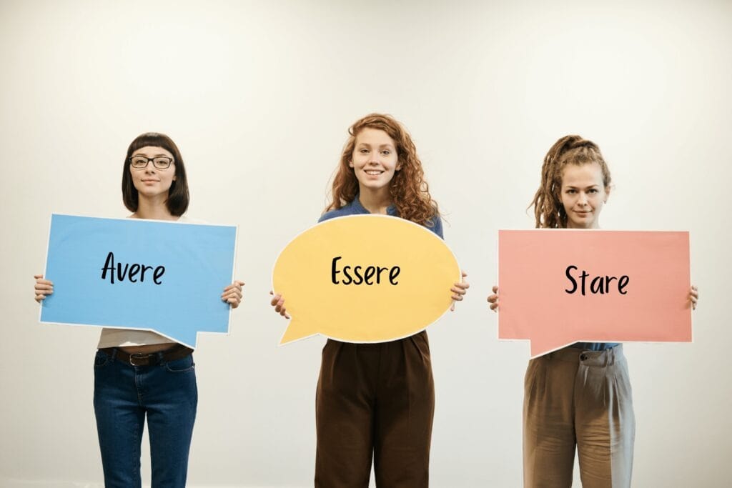 three-women-holding-up-differently-shaped-speech-balloon-cutouts-with-the-words-avere-essere-and-stare-on-them