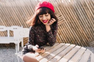 woman in red hat sitting at a wooden table smiling at her phone