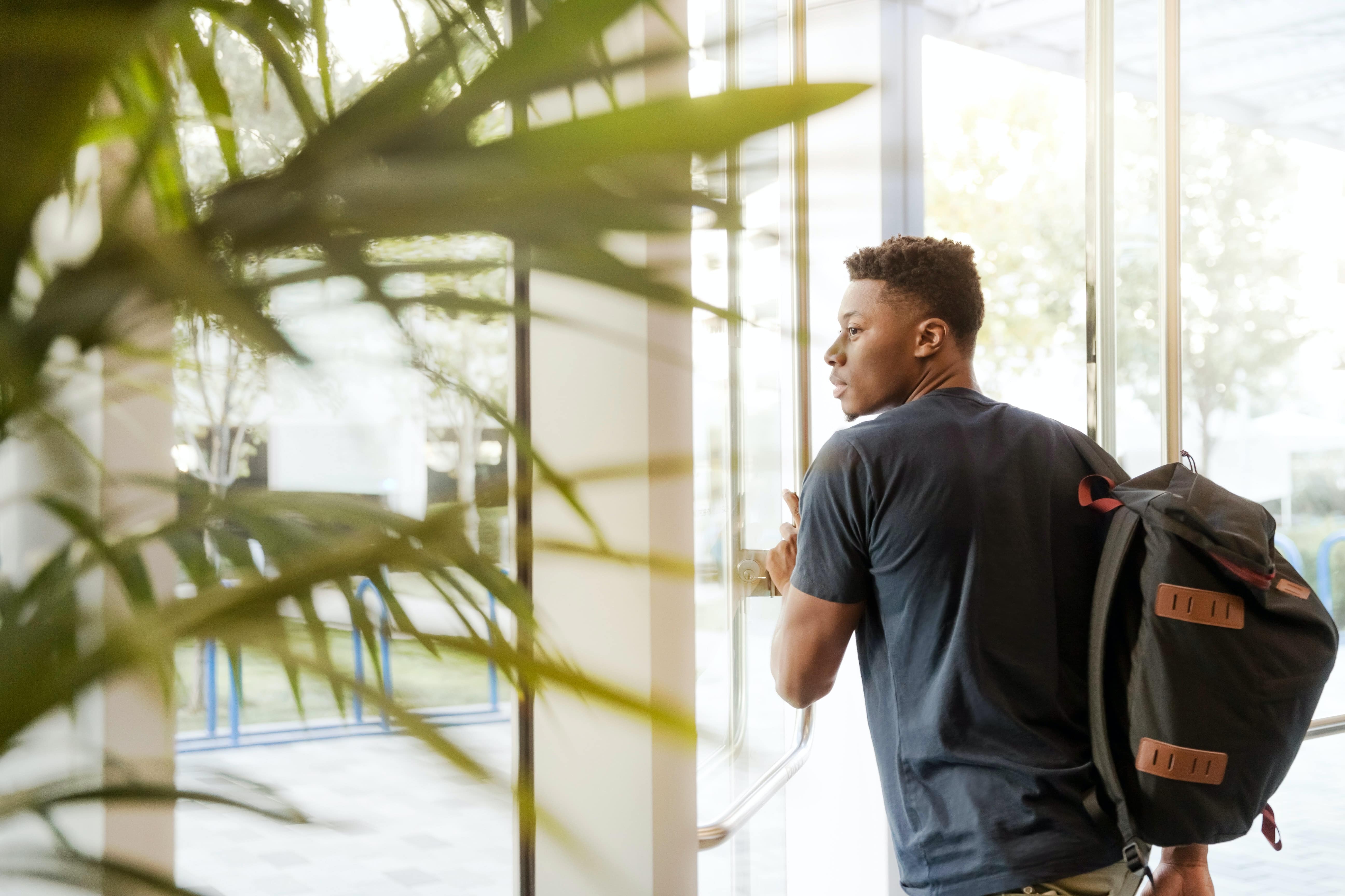 University-aged student looking outside and pushing a glass door to exit a building.