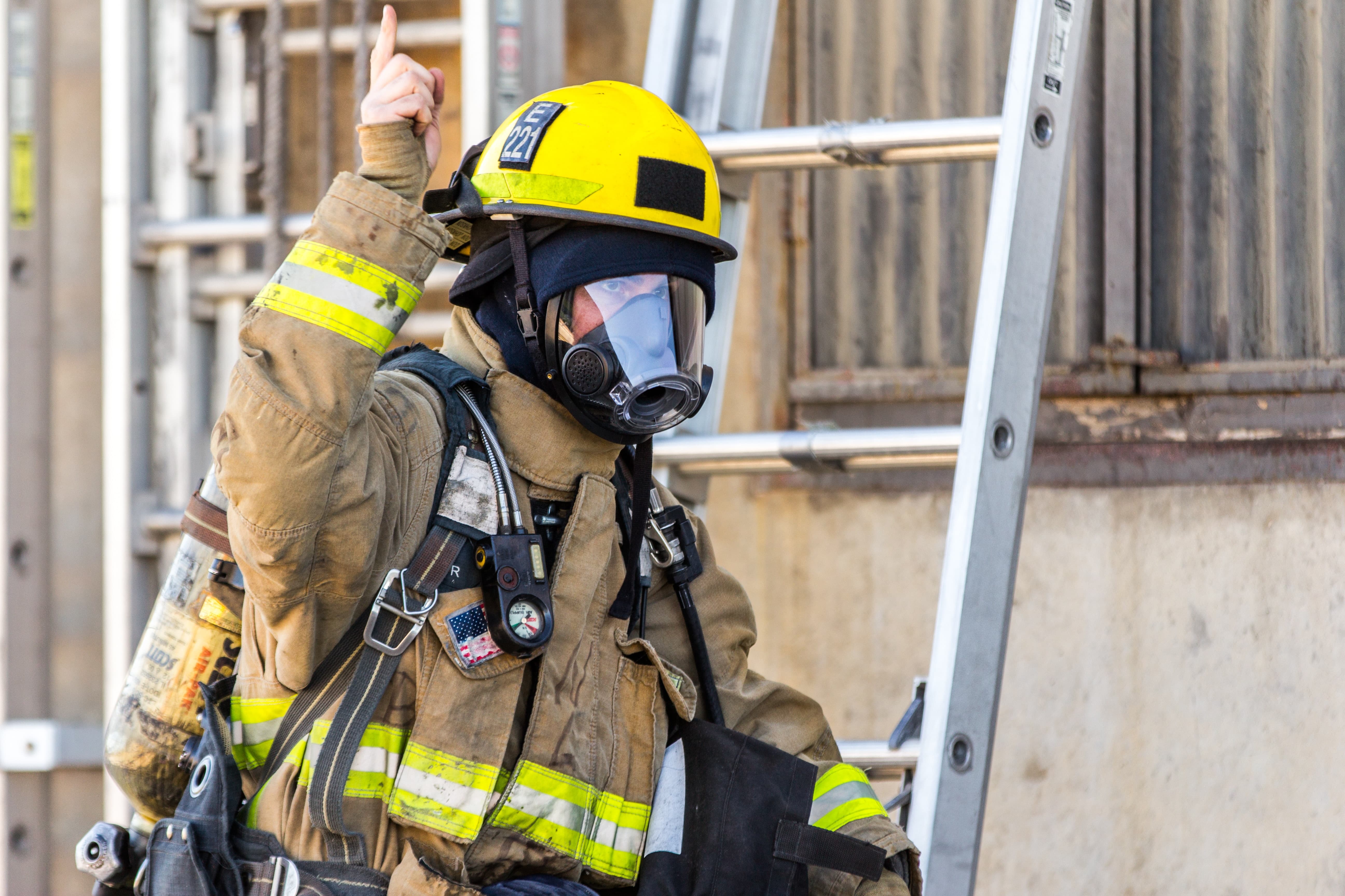 A firefighter wears protective gear and gestures near a ladder.