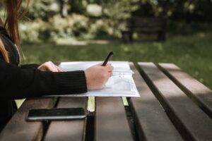 person writing in their notebook on a wooden table at a park