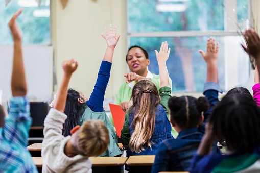 An African-American teacher, a mature woman in her 40s, sitting in front of her class of elementary school students, 6 and 7 years old, reading a book. She has asked a question and all of the children are raising their hands. They are in first grade or second grade. The focus is on two of the girls.