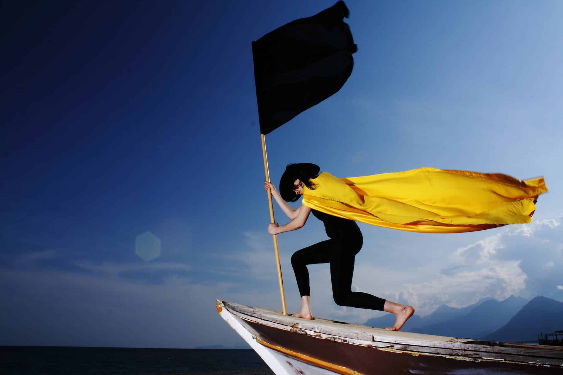 woman-wearing-black-tightfitting-clothes-and-yellow-cape-holding-black-flag-on-edge-of-boat